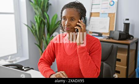 A professional african woman in a red sweater converses on the phone in a modern office environment. Stock Photo