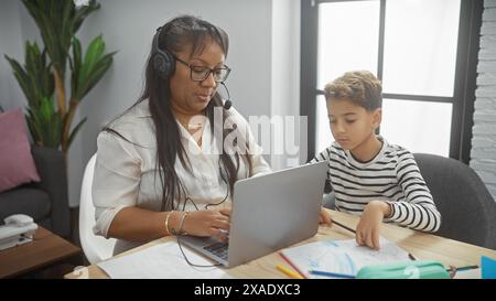 A focused woman with headphones using a laptop and her son drawing next to her in a cozy living room setting. Stock Photo