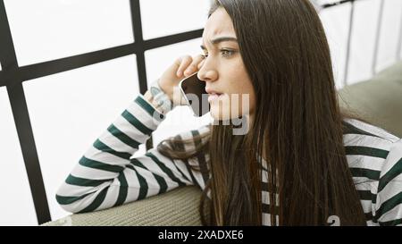 A contemplative young woman on a phone call indoors, exuding a look of concern or deep thought. Stock Photo