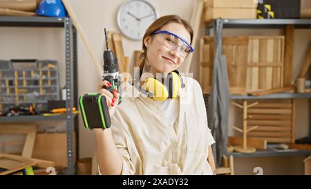A confident woman with a drill stands in a carpentry workshop, showcasing a blend of femininity and craftsmanship. Stock Photo