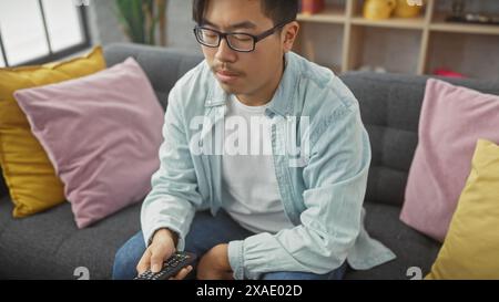 A young asian man sits on a grey couch in a cozy apartment living room, holding a remote control. Stock Photo
