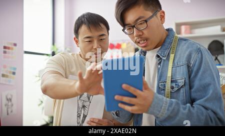 Two asian men collaborating in a tailor shop with one holding a tablet, both focused on design details. Stock Photo