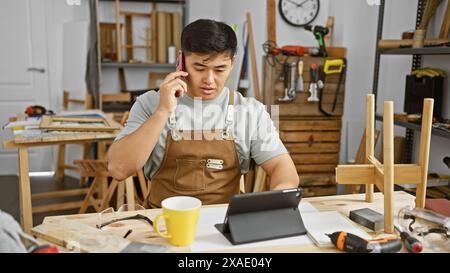 Chinese man in apron talks on phone at carpentry workshop with tools and tablet Stock Photo