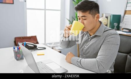 A young asian man enjoying a coffee break at his modern office desk, surrounded by work essentials Stock Photo