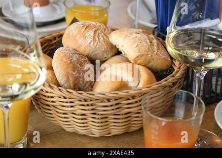 A basket of bread and a glass of orange juice sit on a table. The basket of bread is filled with various types of bread, including rolls and bagels Stock Photo