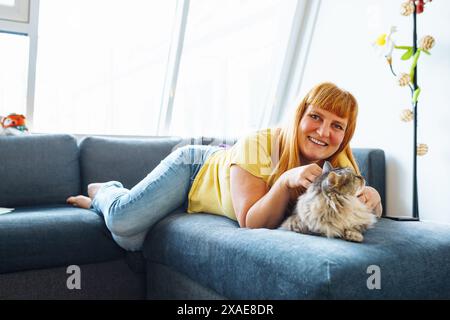 portrait positive blonde middle-aged woman lying on sofa hugging cat taking selfie Stock Photo