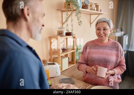 Waist up portrait of smiling senior woman enjoying morning at home in retirement and talking to husband copy space Stock Photo