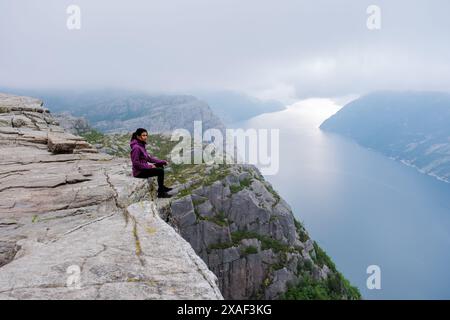 A Asian woman sits on the edge of the Preikestolen cliff in Norway, overlooking a stunning fjord. The misty morning adds to the dramatic landscape. Stock Photo