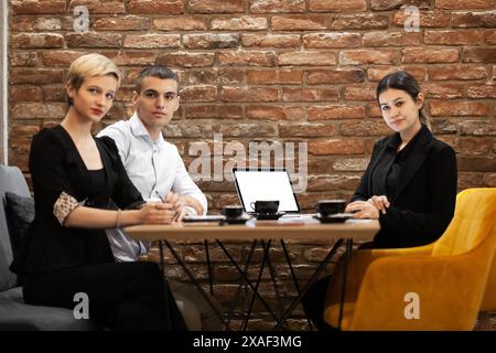 Three young professionals engaged in a discussion at a stylish brick-walled office. Equipped with laptops and coffee, they portray a modern workplace. Stock Photo