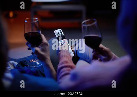 Top view closeup of unrecognizable senior couple watching TV together with hands holding remote control and wine glasses copy space Stock Photo