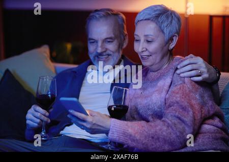Portrait of smiling senior couple using smartphone sitting on couch in blue light and watching TV Stock Photo