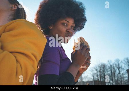 Group of teenagers sitting outside on a basketball field with stands eating kebab, playing basketball, having fun together, listening to the music. Stock Photo