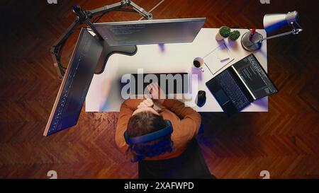 Top down view of frenzied developer suffering from adhd, impatiently arranging monitors while writing code. Jittery IT admin doing multitasking in office, drinking coffee, listening music, aerial shot Stock Photo