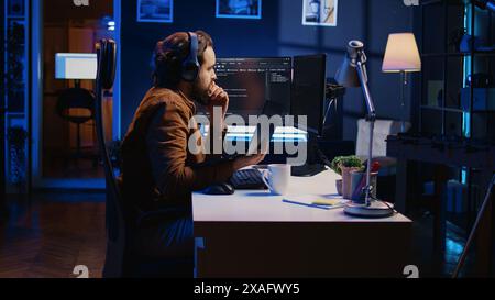 Engineer typing code in apartment office, using javascript programming language to develop software application. It admin using multimonitor desktop setup and notebook to finish tasks, camera B Stock Photo