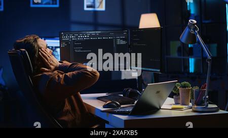 Fatigued software programmer working overnight at home, taking break from coding to stretch at desk chair. Overworked IT professional feeling exhausted, pausing tasks to rest, camera A Stock Photo