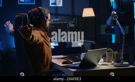 Software programmer taking break from coding, putting headphones on and stretching on desk chair during remote job shift. IT professional pausing tasks, listening music, loosening up, camera A Stock Photo