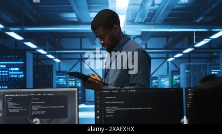 Manager uses tablet, supervising team programming servers providing computing resources for different workloads. Inspector using device to oversee engineers in data center mending supercomputers Stock Photo