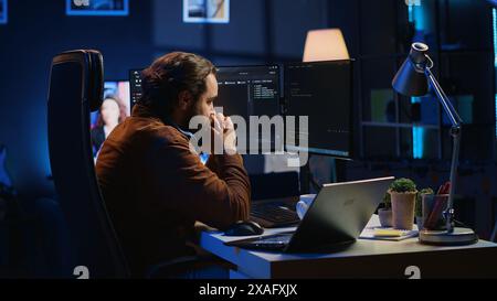 Technician focused on typing complex code in neon lit home office, developing software application. It specialist removing headphones to better concentrate on difficult task, camera A Stock Photo