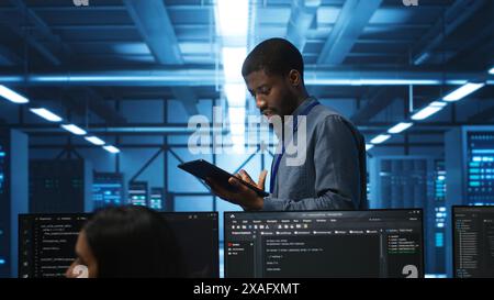 Engineer inputting data on tablet in server hub, monitoring data center energy consumption across hardware. Technician inspecting mainframes, ensuring malfunctions are repaired Stock Photo