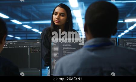 Manager in data center talking with employees about annual system review after checking parameters on laptop. Supervisor overseeing server clusters discussing with multiethnic team Stock Photo