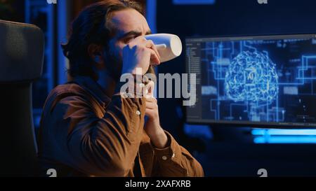 Computer scientist updating artificial intelligence neural networks, drinking coffee and writing code scripts in personal office. IT expert enjoying hot beverage while running AI simulations, camera A Stock Photo