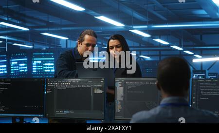 Multiethnic engineering team in server hub monitoring data center energy consumption across hardware using laptop. Multiracial colleagues inspecting supercomputers, ensuring malfunctions are fixed Stock Photo