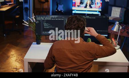 Computer scientist at home office desk writes binary code scripts and drinks from mug of coffee. IT expert in apartment enjoying tea while using programming to safeguard consumer data, camera B Stock Photo