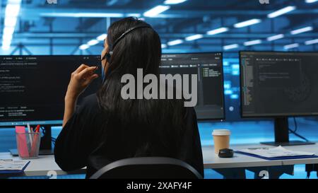 Computer scientist upgrading equipment in data center, wearing headphones to listen music. Programmer at PC desk in high tech establishment overseeing server rows, typing on keyboard Stock Photo