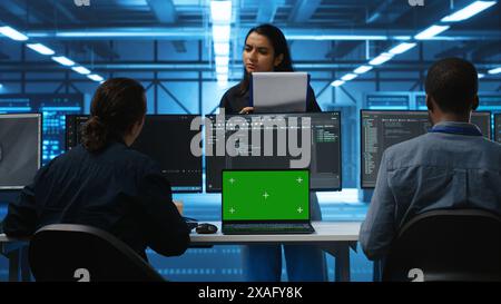 Multiethnic engineers working in high tech server hub, using green screen laptop and PCs to analyze data. Diverse employees examining infrastructure in data center, using chroma key notebook Stock Photo