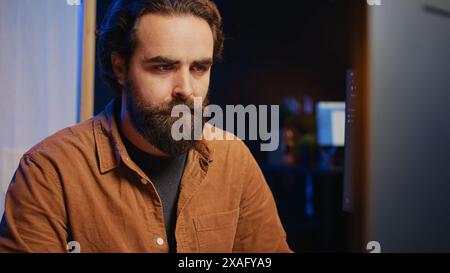 Software technician in apartment office deeply immersed in coding on computer at desk, close up. IT freelancer absorbed by work, focused on screen, writing lines of code at workstation, camera B Stock Photo