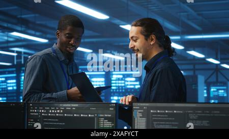 Coworking system administrators in server hub monitoring data center energy consumption across hardware using tablet. Colleagues inspecting supercomputers, ensuring malfunctions are fixed Stock Photo