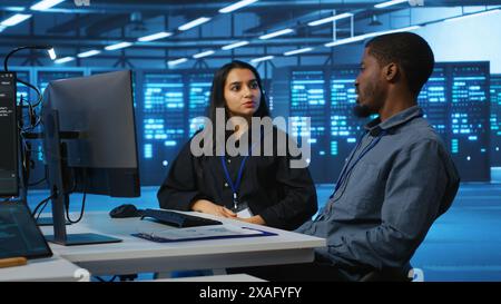 Multiracial team of IT engineers in server room brainstorming ways to fix equipment storing datasets. Indian and african american coworkers discussing how to mend racks doing computational operations Stock Photo