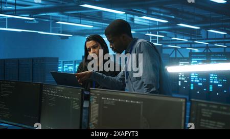 Coworking technicians looking at laptop screen, working in data center, ensuring optimal performance. Colleagues monitoring energy consumption across server rigs components, doing brainstorming Stock Photo