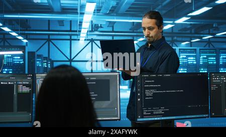 Engineer typing on laptop in server hub, checking recovery plan, monitoring data center energy consumption across components. Technician inspecting rackmounts, doing routine disk checks Stock Photo