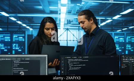 Technicians working in high tech server hub, analyzing data on laptop to ensure seamless data flow. Coworkers examining equipment in data center, ensuring system integrity and security Stock Photo
