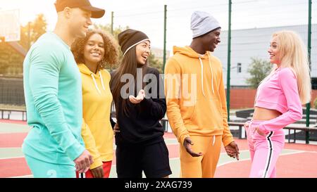 Group of young sportspeople of different nationalities in colorful sportswear talking at university stadium. Multi-ethnic group teenage friends spending time together at sportsground. Stock Photo