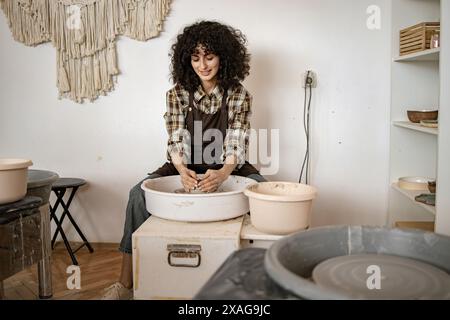 Smiling young female potter shaping clay on a pottery wheel in her studio, focused on creating ceramic dishes. Stock Photo