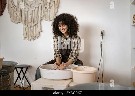 Female potter using wheel to sculpt ceramic bowl in cozy studio. Curly haired woman enjoying creative process. Stock Photo