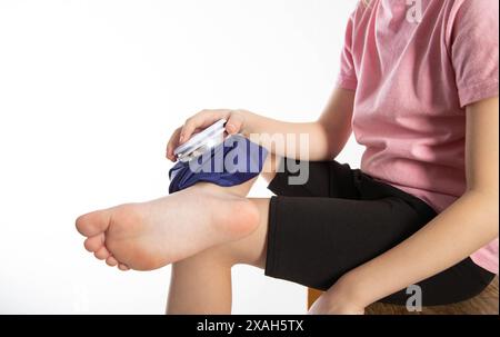 A girl applies a blue medical bag with ice to her ankle joint for a bruise on her leg. Local cryotherapy. Pain relief with cold, close-up. Copy space Stock Photo