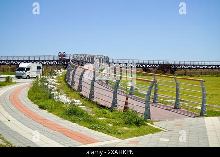 Samcheok City, South Korea - May 18th, 2024: A curved walkway at Isabu Plaza leads towards a wooden bridge and a scenic view of the area. Vehicles and Stock Photo