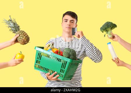 Thoughtful young man holding full shopping basket with credit card and food on yellow background Stock Photo
