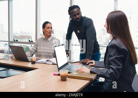 Three young people are sitting with laptops and a book, arguing among themselves about a business. A man is standing looking at the woman using the la Stock Photo