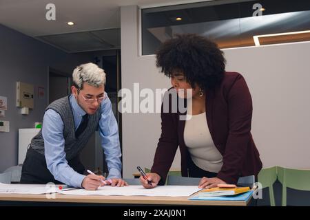 Two young people woman and man, are working together on a project they are talking and taking notes on a paper. They are writing on a table with pens Stock Photo