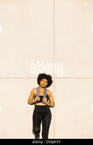 Woman leaning against a wall with a towel around her neck. Portrait of a young woman taking a break after her workout. Stock Photo