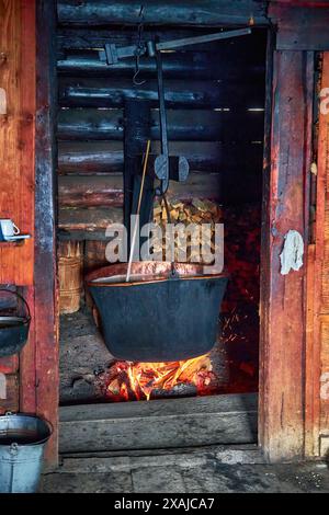 A large vat in which cheese is cooked over an open fire. The black cauldron is prepared over an open wood fire. Cheese factory in the mountains. Stock Photo