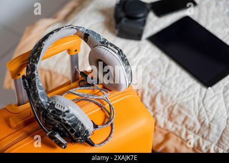Close-up of a pair of headphones resting on an orange suitcase with a camera, smart phone, and tablet in the background on a bed. Stock Photo