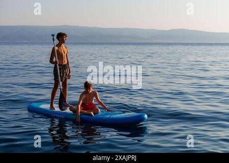 Two teenage boys explore the coastline on a stand up paddle board, as the sun sets in Omis, Croatia.There are silhouetted mountains in the background. Stock Photo