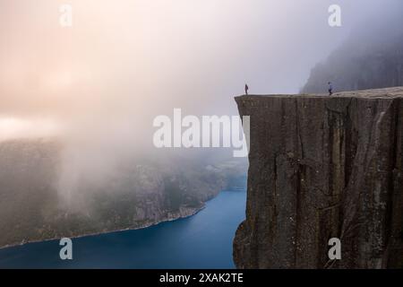 A misty morning scene in Norway. Two figures stand on the edge of Preikestolen, a famous cliff face, with a view over a valley and a deep blue fjord. men and women hiking the Preikestolen, Norway Stock Photo