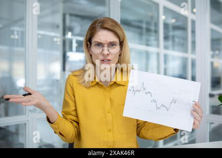 Businesswoman expressing confusion while analyzing a stock market graph during a meeting in a modern office setting. Stock Photo