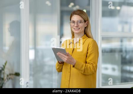 Confident young professional woman with glasses holding a tablet and smiling in a modern office environment. Stock Photo
